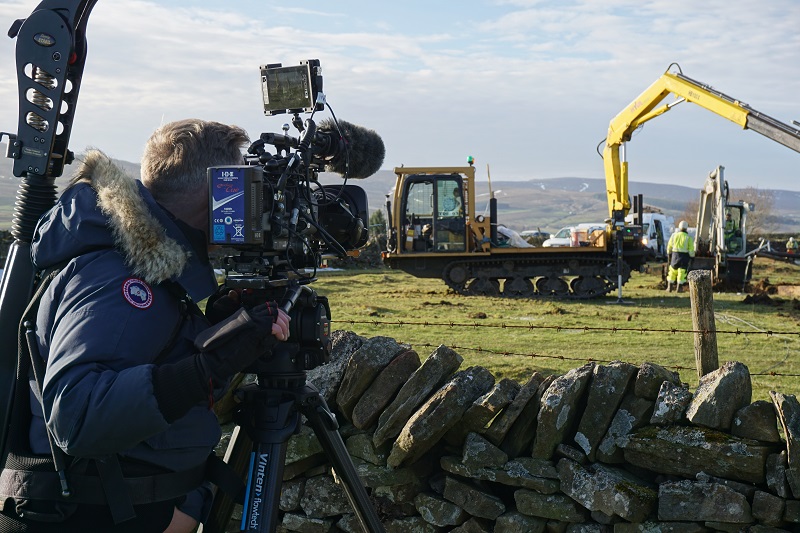 ross marshall with a large video camera leaning on a stone wall filming large plant machinery at work in a field